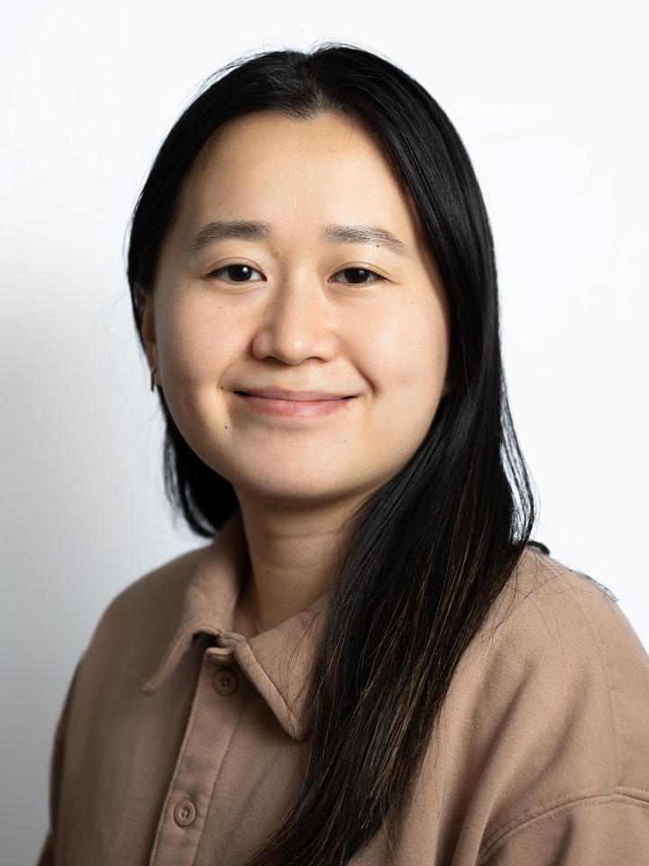 A portrait of Shiya, a Chinese person with long black hair and wearing a brown collared shirt, looking into the camera and smiling. Posing in front of a blank white background. 