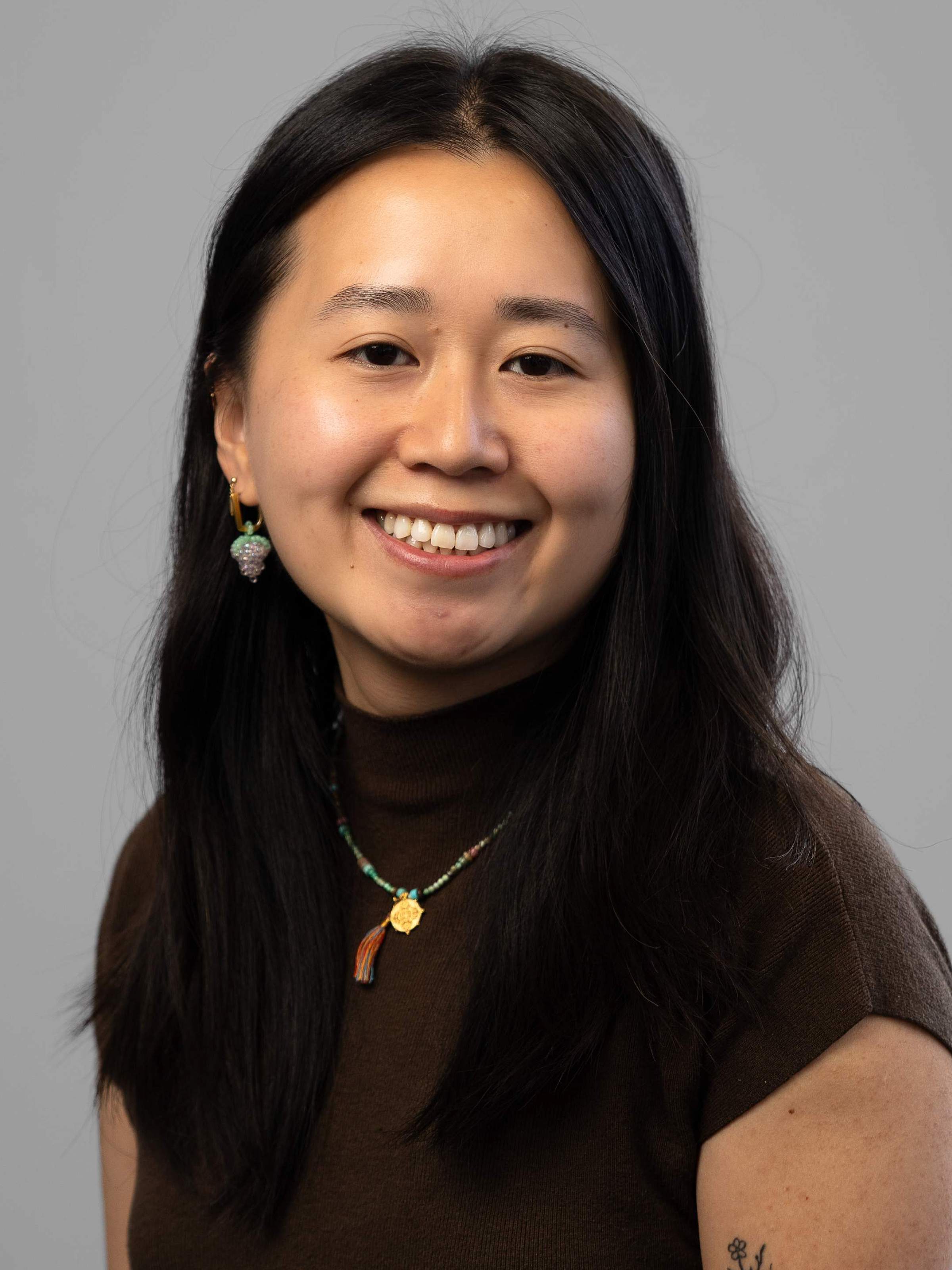 A portrait of Shiya, a Chinese person with long black hair and wearing a brown collared shirt, looking into the camera and smiling. Posing in front of a blank white background. 