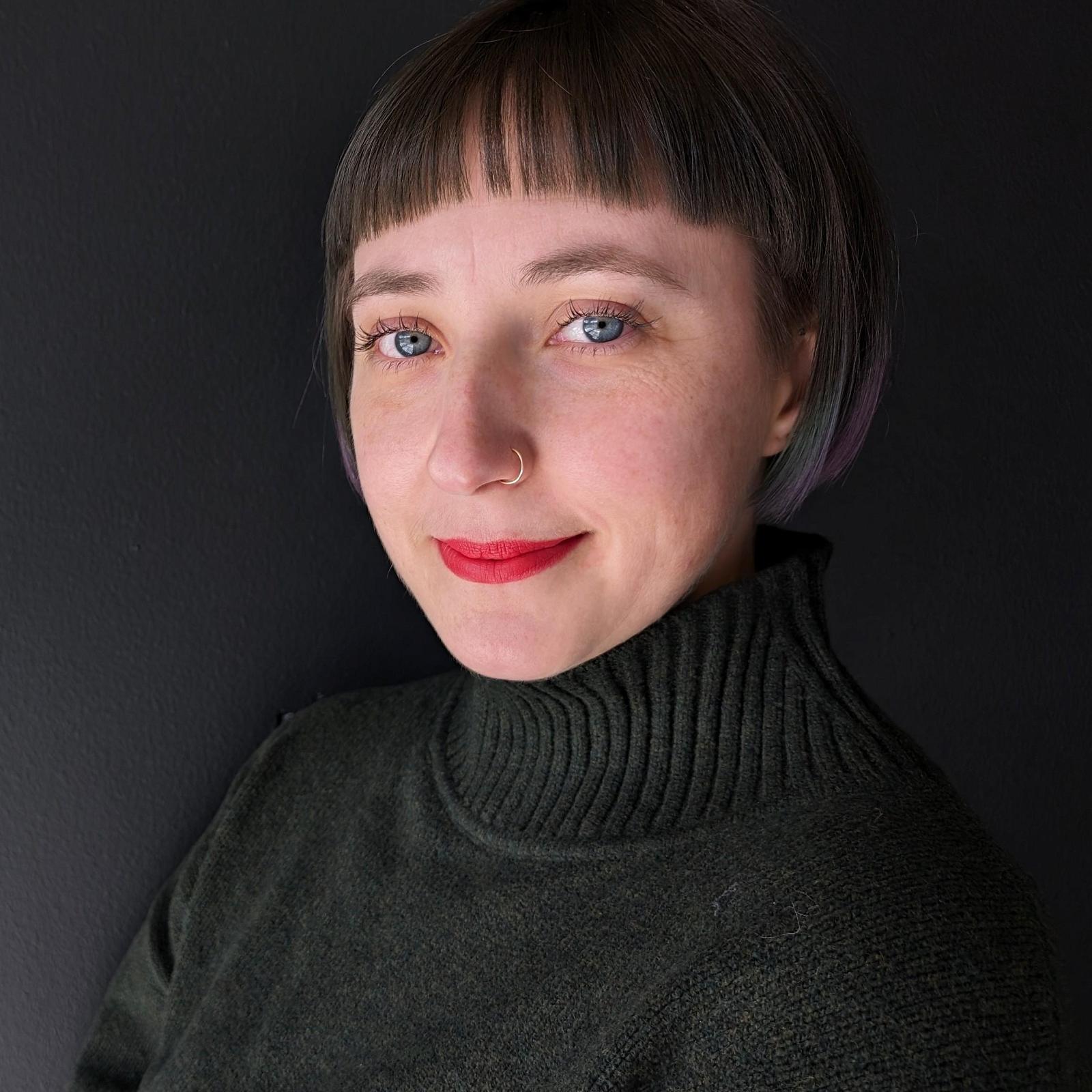 A person with blue eyes, short brown/blue/purple hair with bangs smiles at the camera wearing red lipstick. They have on a forest green mock neck sweater. The background is dark grey.