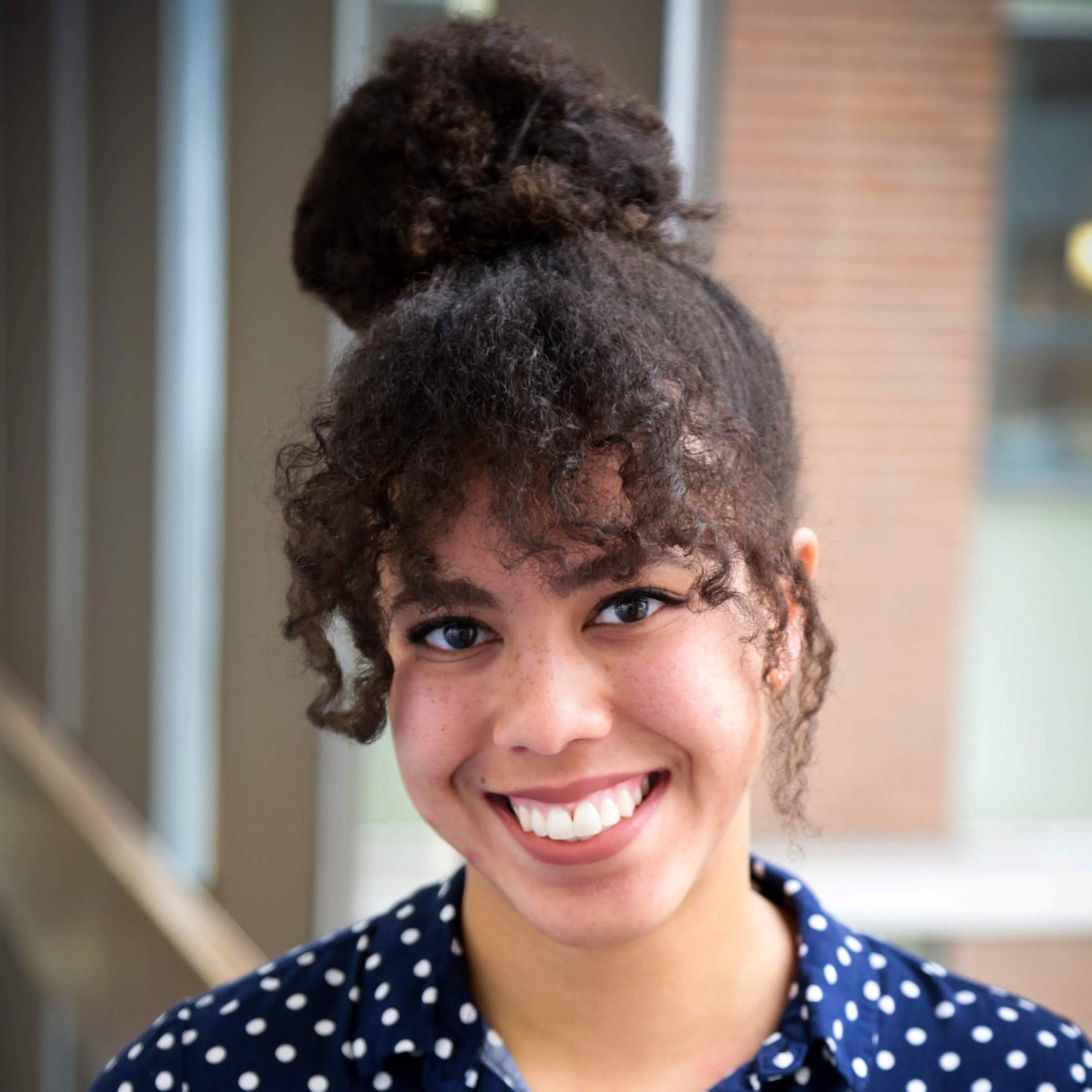 Headshot, Ashley Joseph, curly brown hair in a bun and a navy blue polka dot shirt