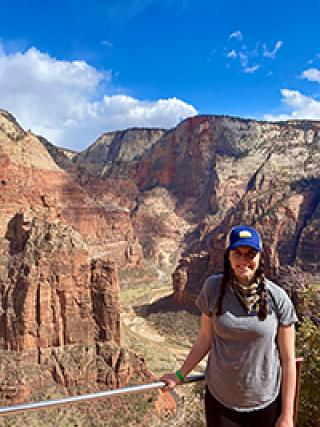 woman in a gray shirt and a blue had with braided hair in front of red rocks