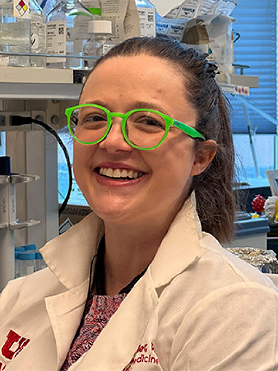 picture of a young woman in a lab with brown hair pulled in a pony tail and a bright green glasses