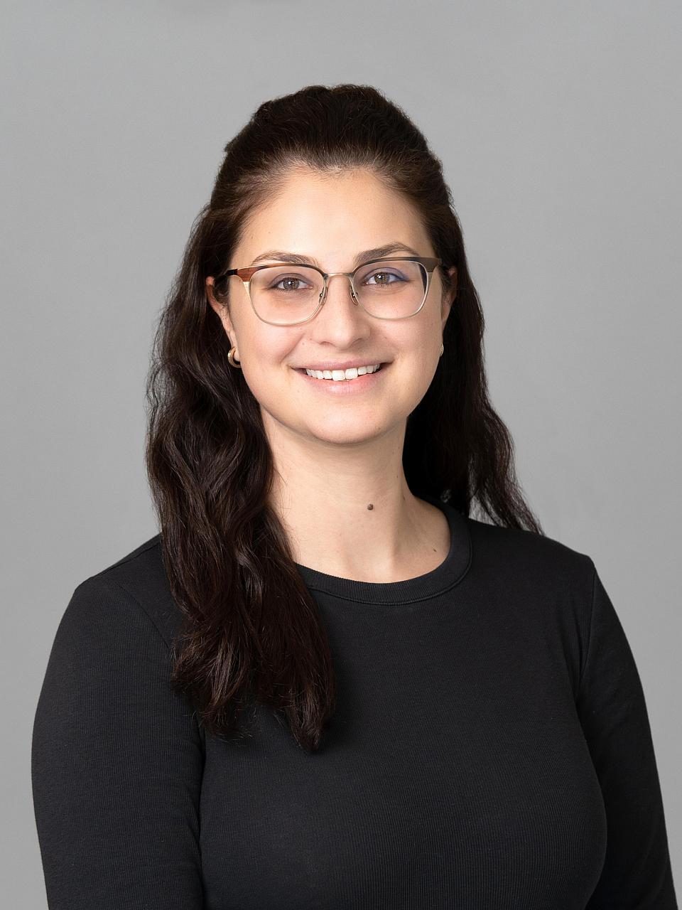 Photo of a woman, Dr. Aleksandra Abrahamowicz, with dark hair and glasses smiling and looking at the camera wearing a black shirt.