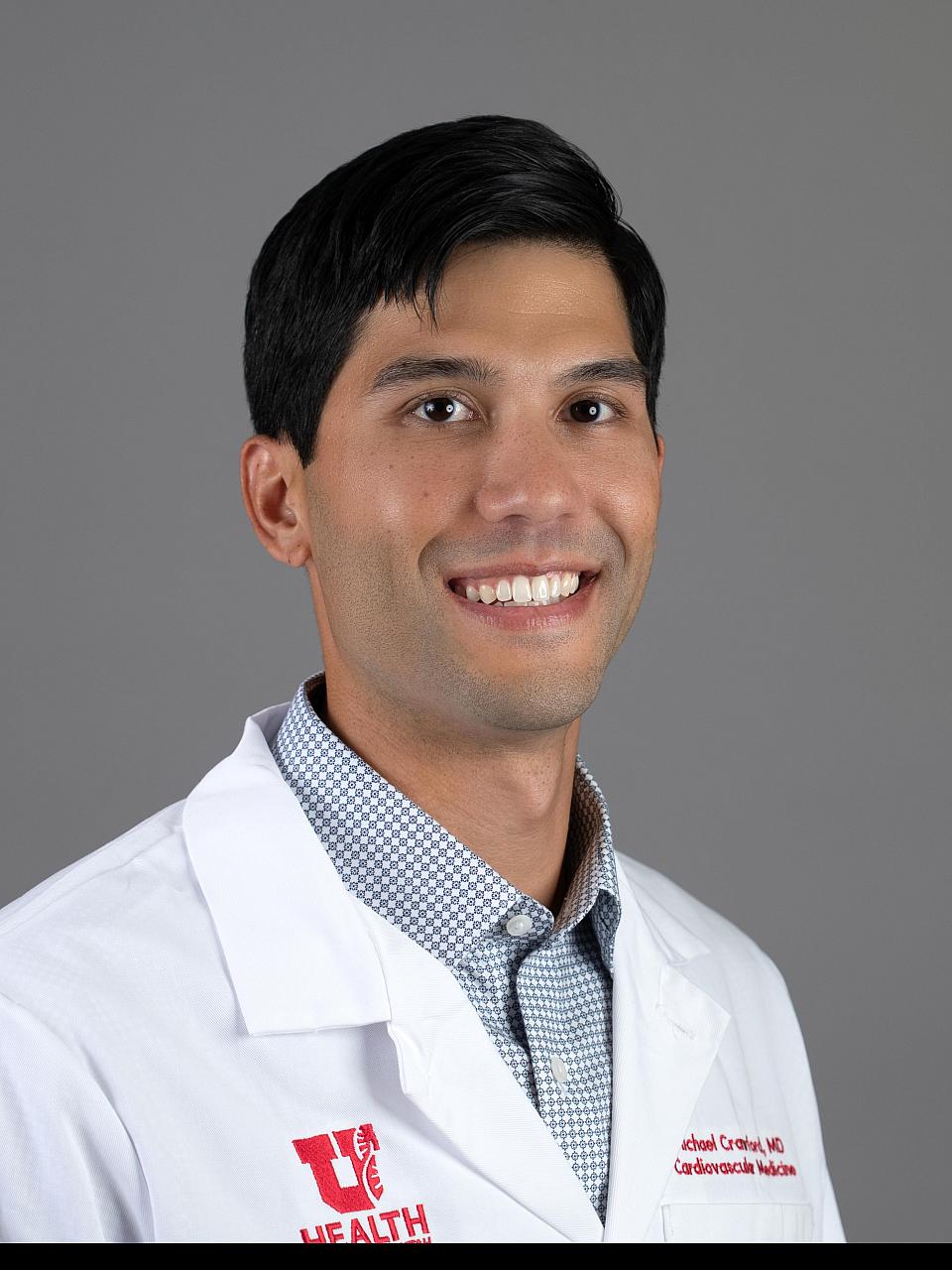 Photo of a man, Dr. Michael Crawford, with short dark hair wearing a checked shirt and white coat smiling at the camera.