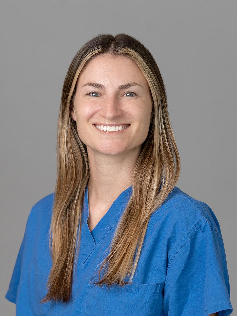 Photo of a woman, Dr. Tally Desmarais, with long light brown hair smiling and looking at the camera wearing blue scrubs.