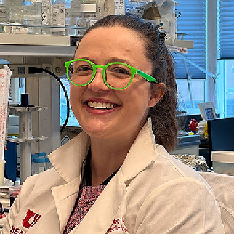 picture of a young woman in a lab with brown hair pulled in a pony tail and a bright green glasses