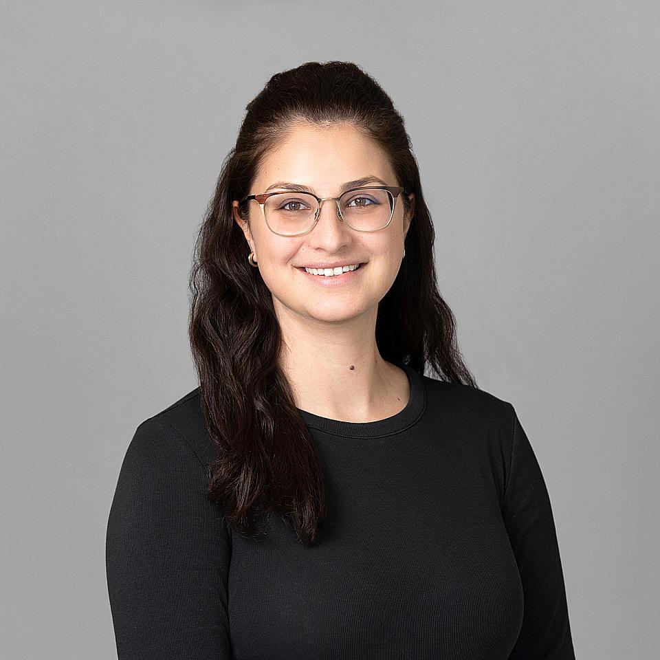 Photo of a woman, Dr. Aleksandra Abrahamowicz, with dark hair and glasses smiling and looking at the camera wearing a black shirt.