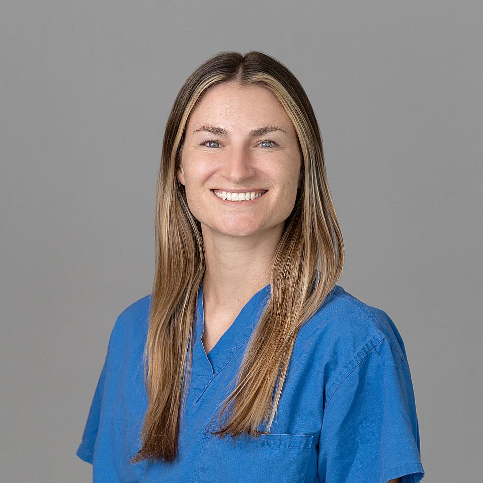 Photo of a woman, Dr. Tally Desmarais, with long light brown hair smiling and looking at the camera wearing blue scrubs.