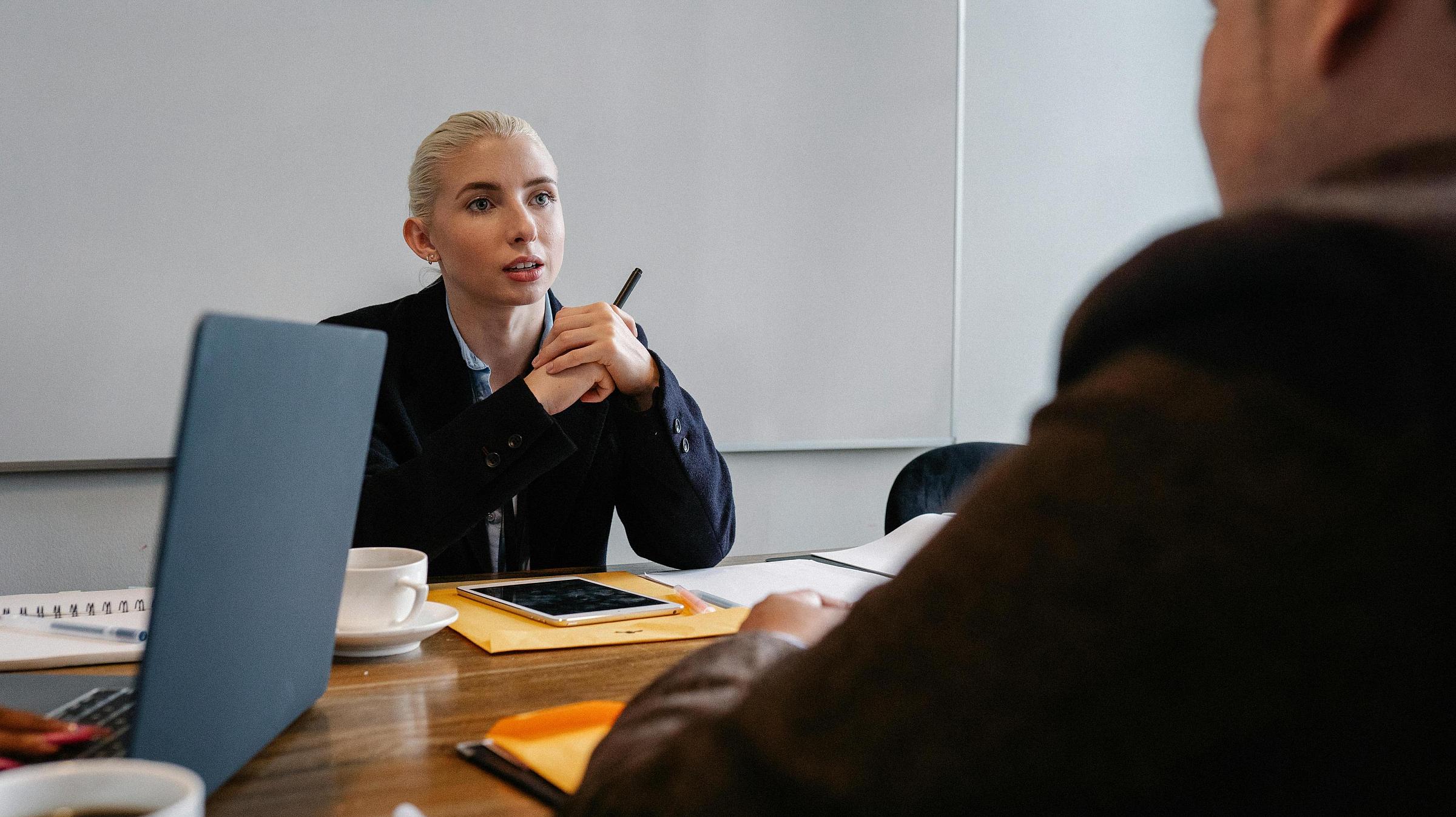 Professional woman in a suit sitting at a table with a pen, laptop, documents, and paperwork talking to another person