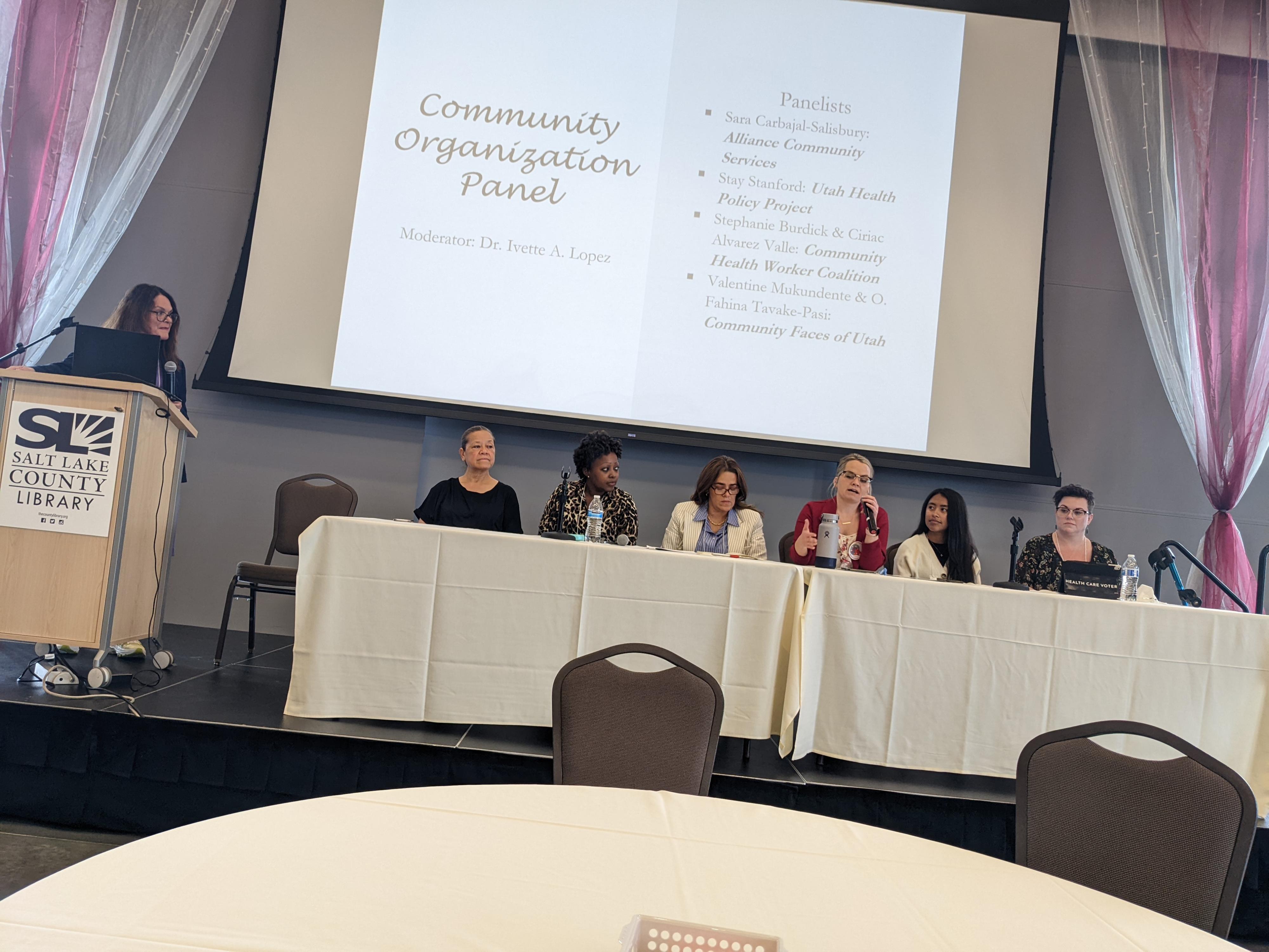 A group of individuals sit at a conference panel with a presentation reading Community Organization Panel above them