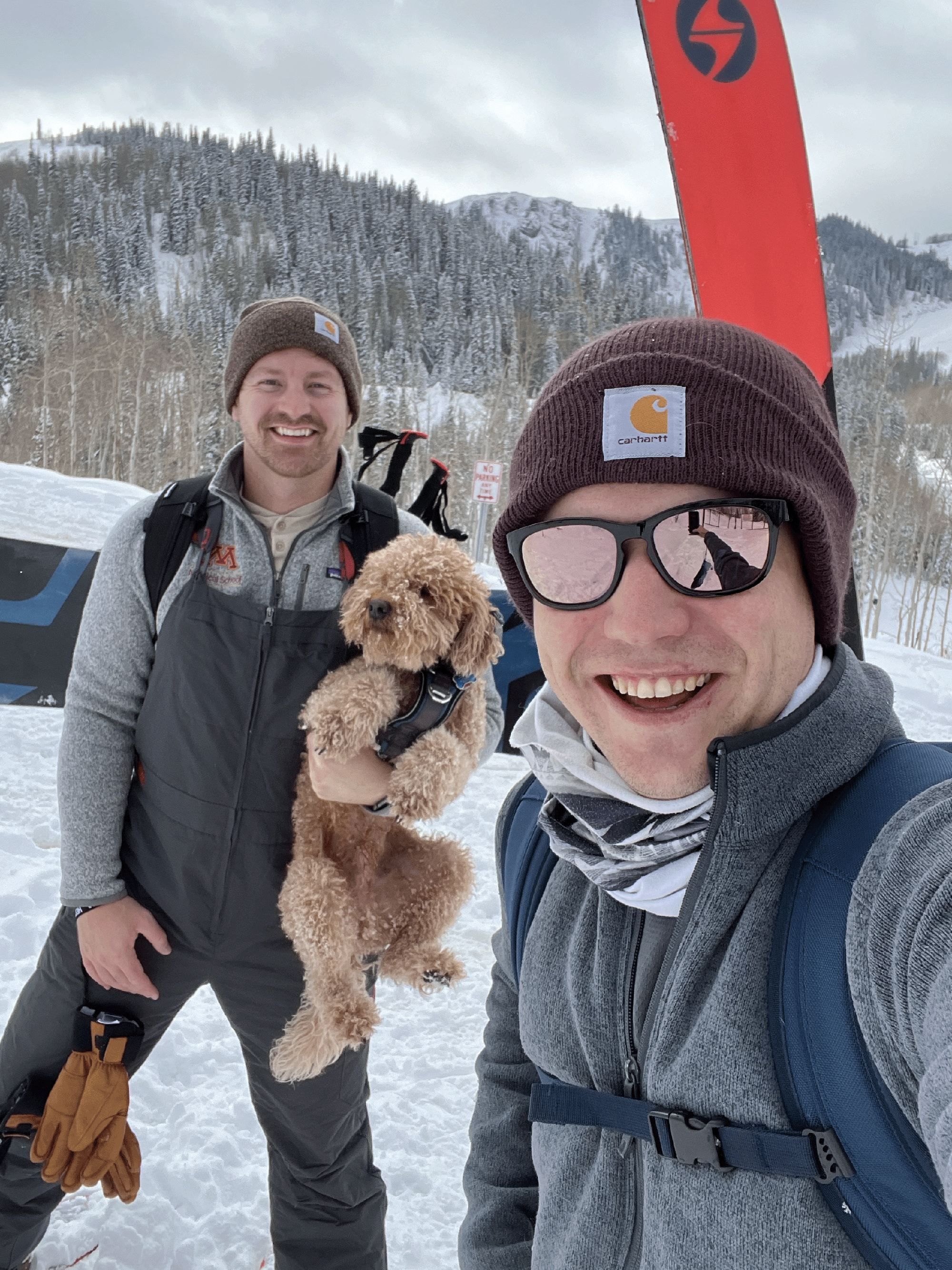 Two people standing in the snow. One has skiis; the other is holding a dog