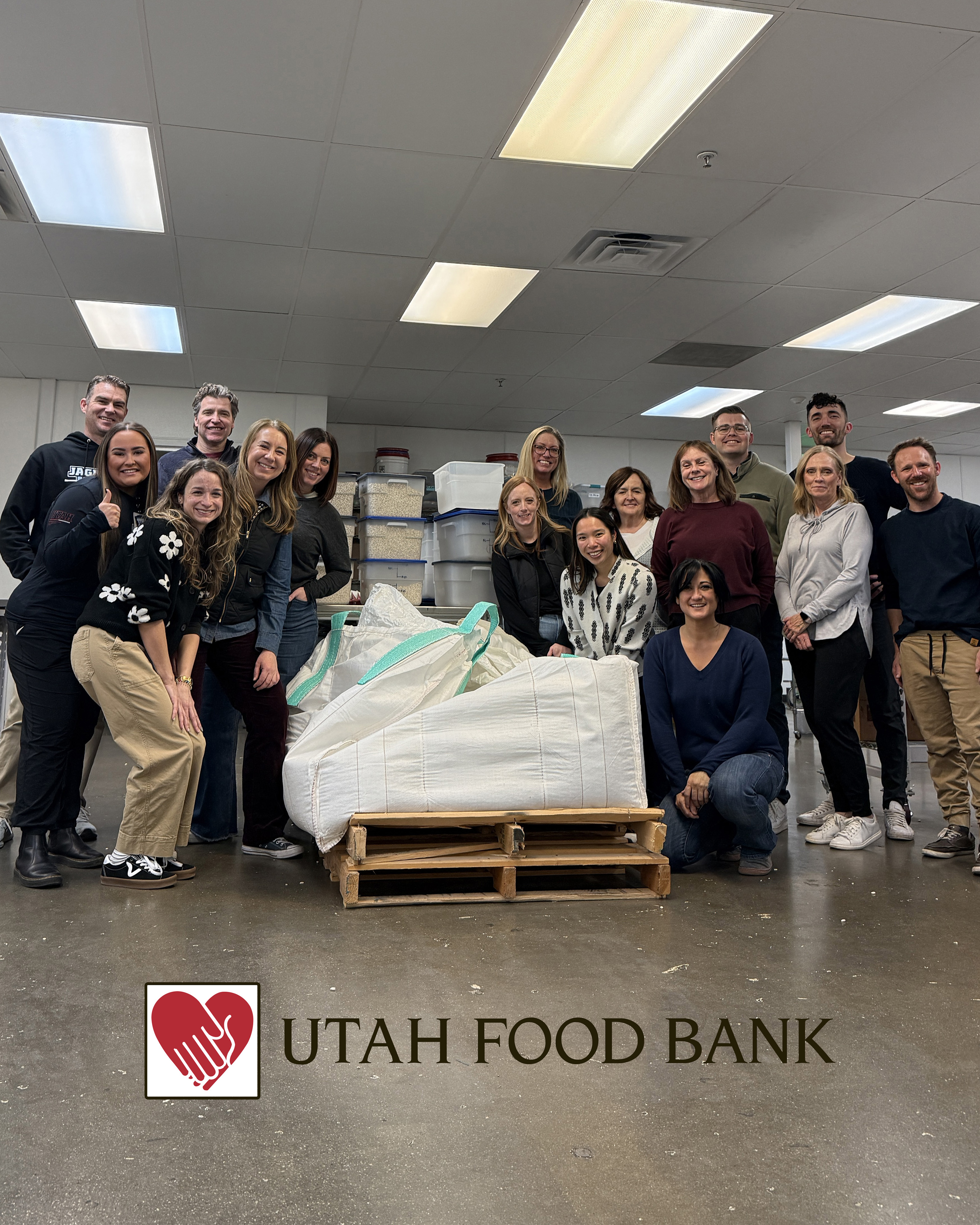 An image of Dept. of Anesthesiology administrative staff at the Utah Food Bank smiling and posing around a bag of beans. At the bottom of the image is the logo for the Utah Food Bank, two hands forming a heart in red on a white background with "Utah Food Bank" in black written next to it.