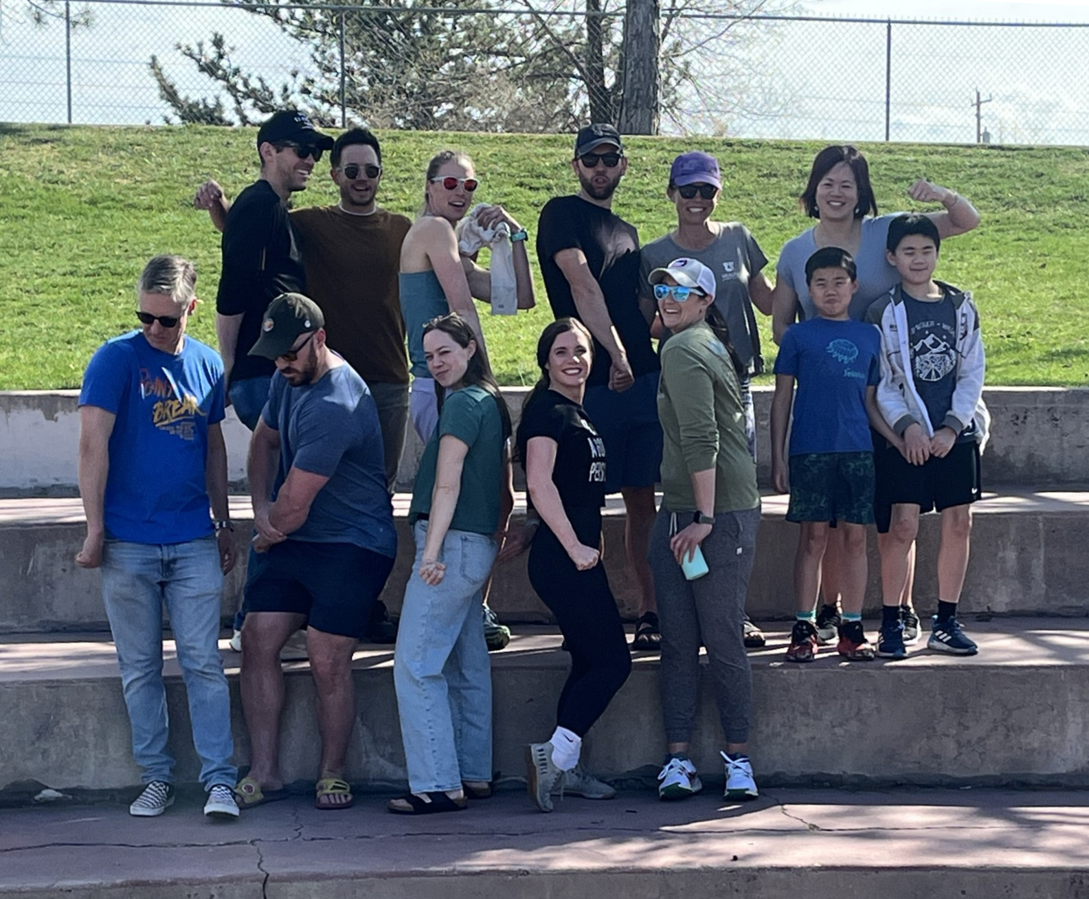 Participants in the 2025 Burpee challenge pose for a group photo on the stone steps of an amphitheater in two rows. The back row includes six adults and two children, and the front row includes five adults. All the adults are flexing their arms.