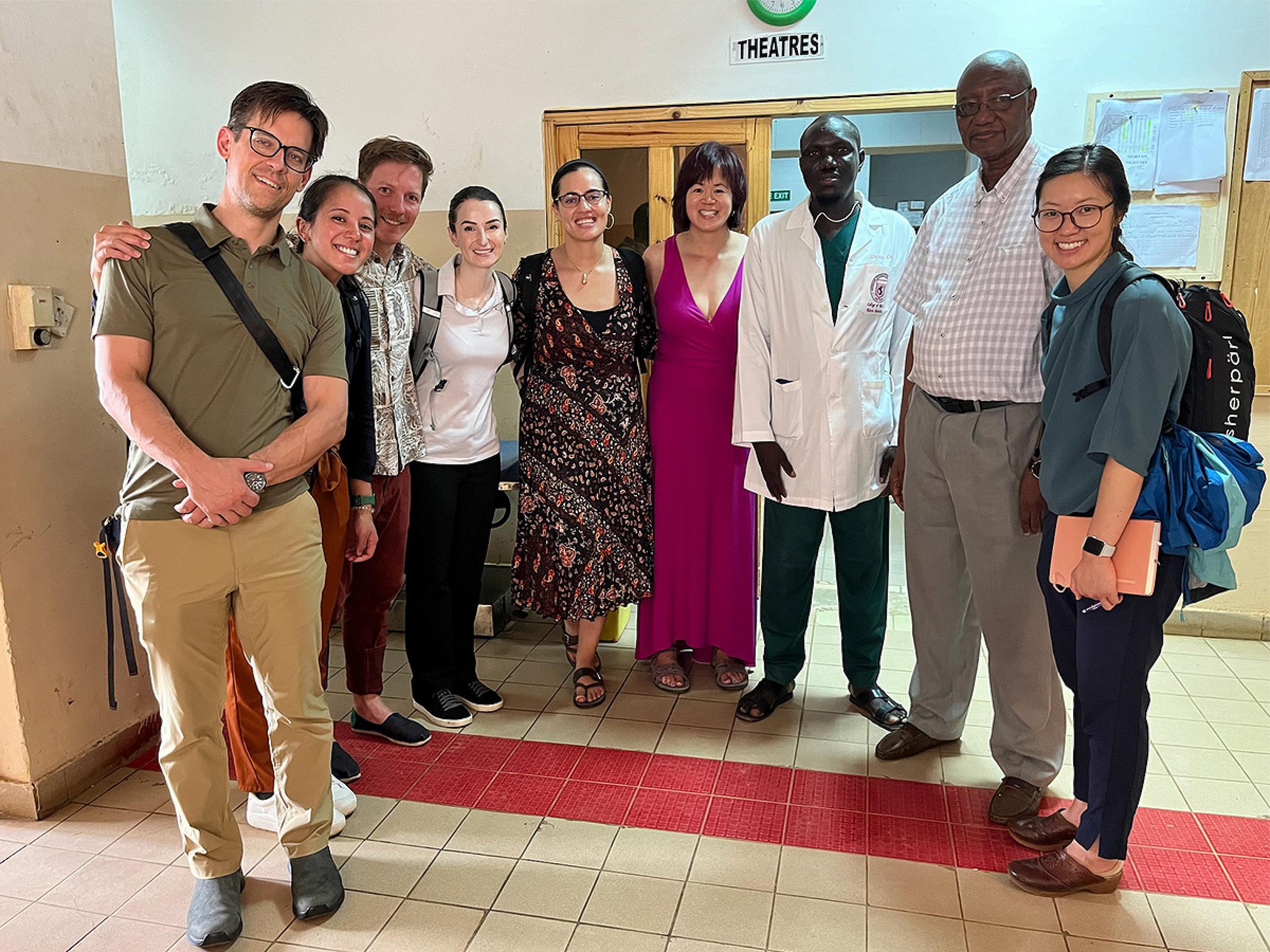 9 people standing together for a group photo in the hallway of a medical facility. 7 are from the University of Utah's Department of Anesthesiology, 2 are medical professionals from the hospital in The Gambia.