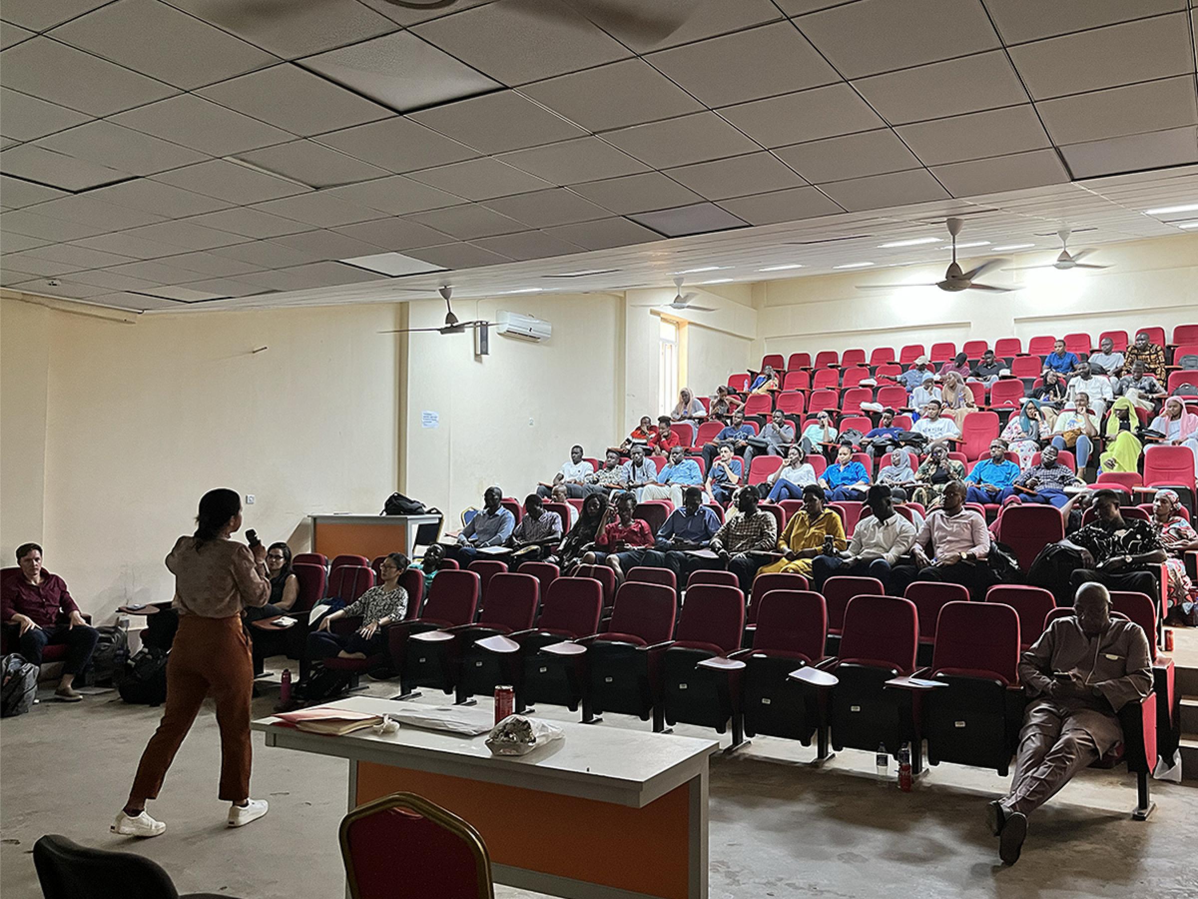 A resident teaching a large group of people in a stadium-seated conference room.