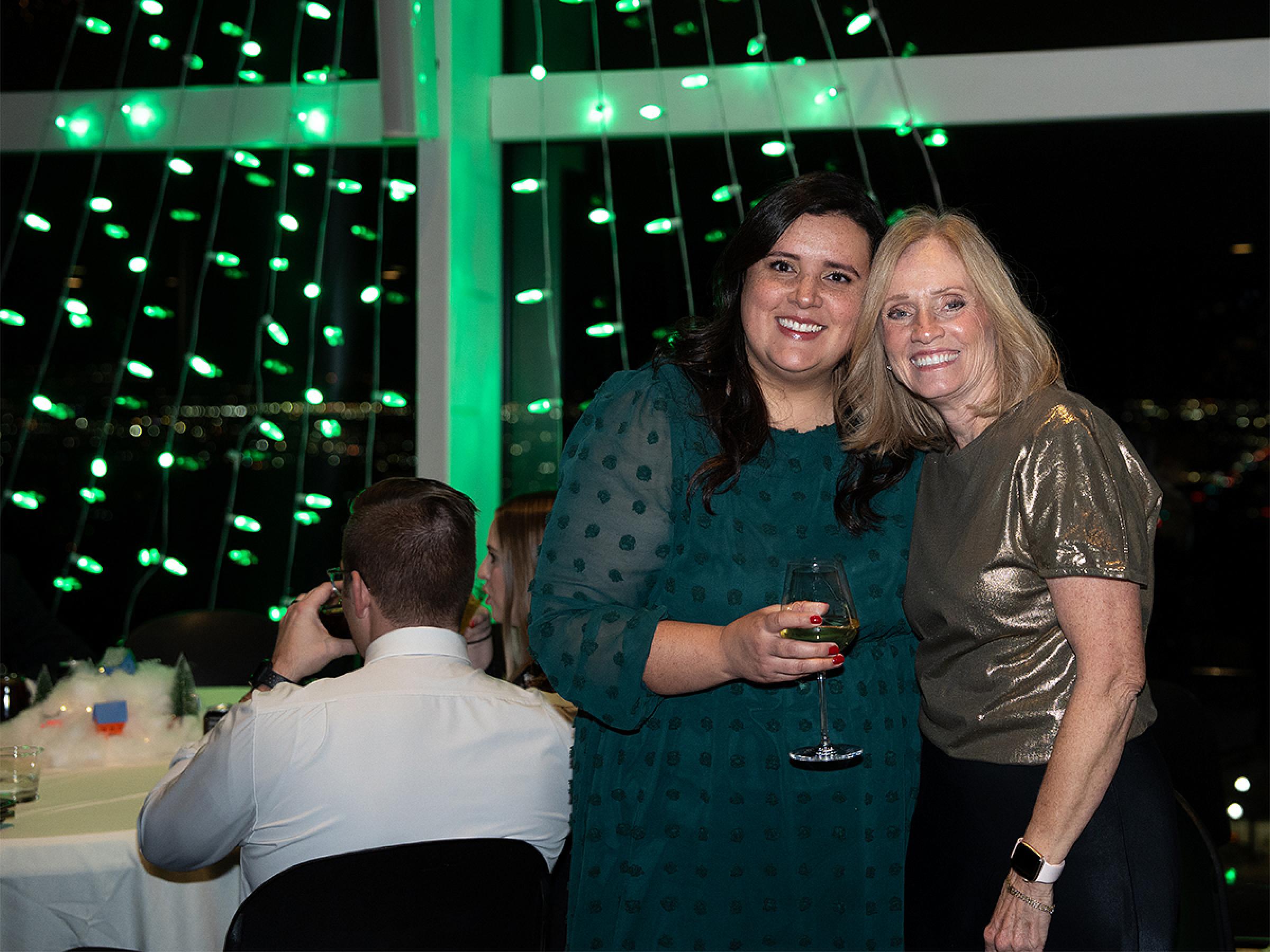 Two people smiling for a photo together in front of a green Christmas tree made of lights. 