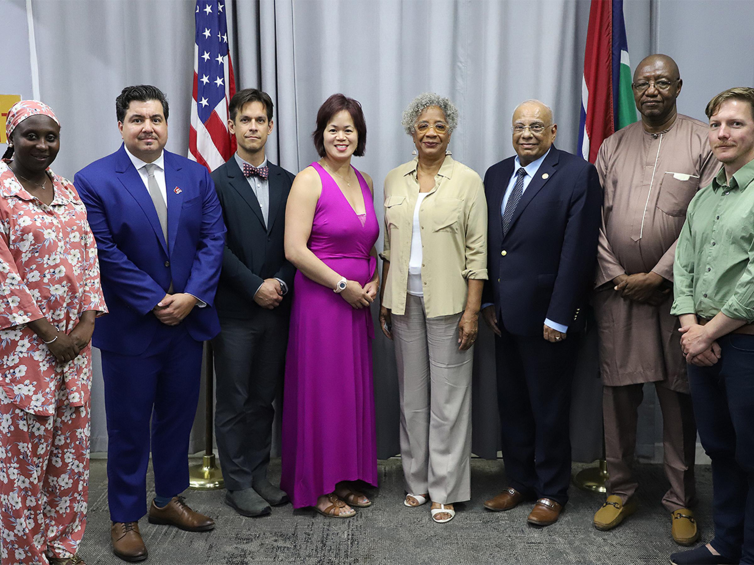 A group of 8 people standing together with the Ambassador of The Gambia in front of the American flag and the flag of The Gambia.