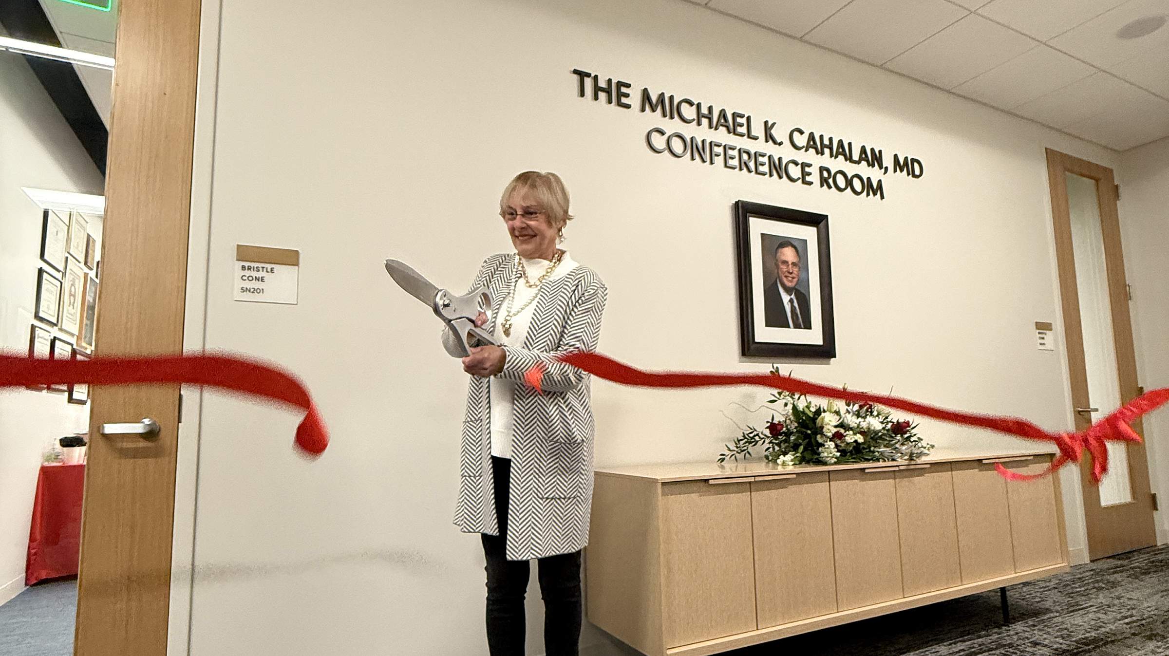 Marianne Cahalan using large scissors to cut the red ribbon in front of the doors to the Michael K Cahalan, MD Conference Room. 