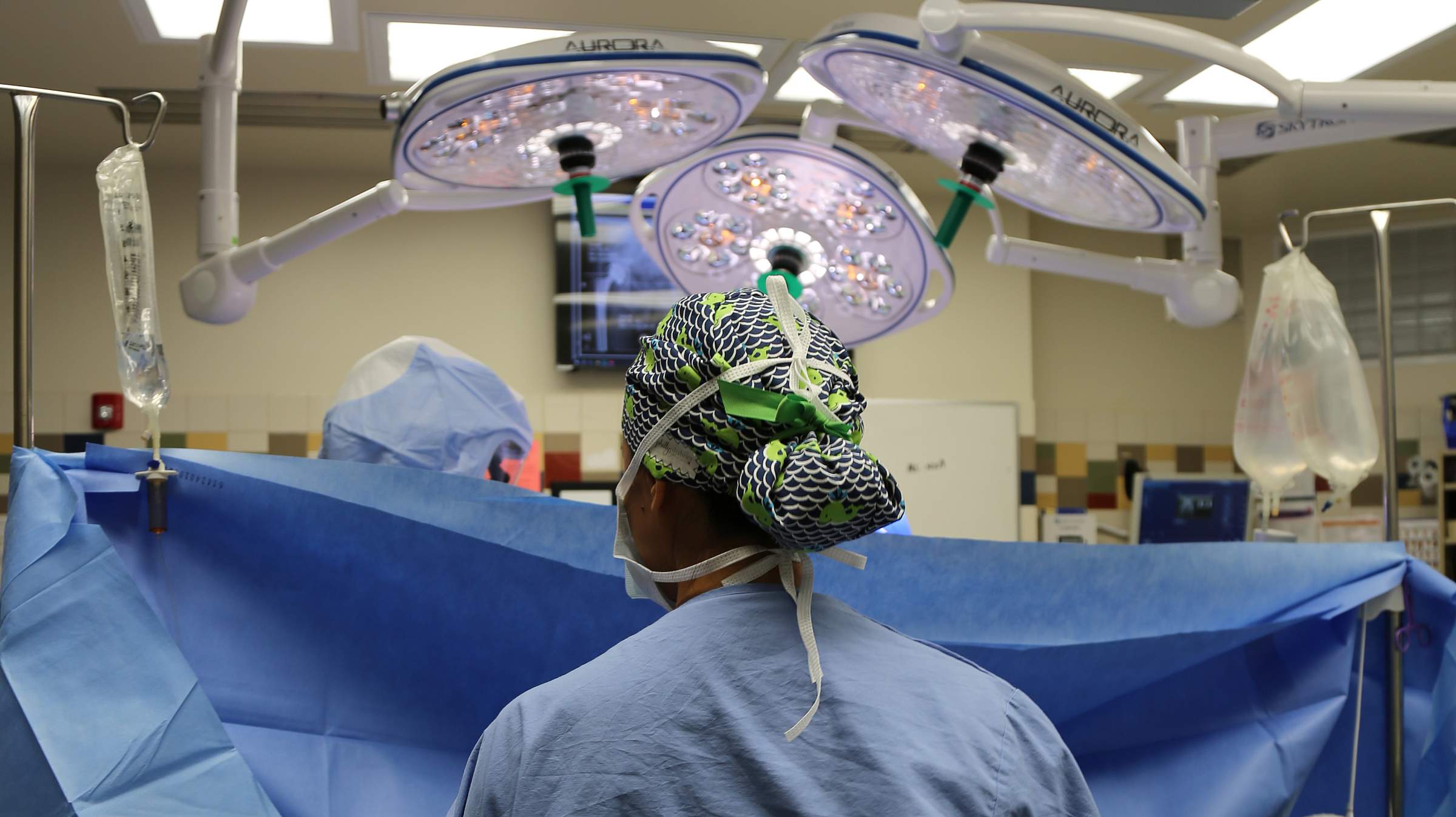 An anesthesiologist at the head of a surgical table in an OR wearing a green scrub cap.