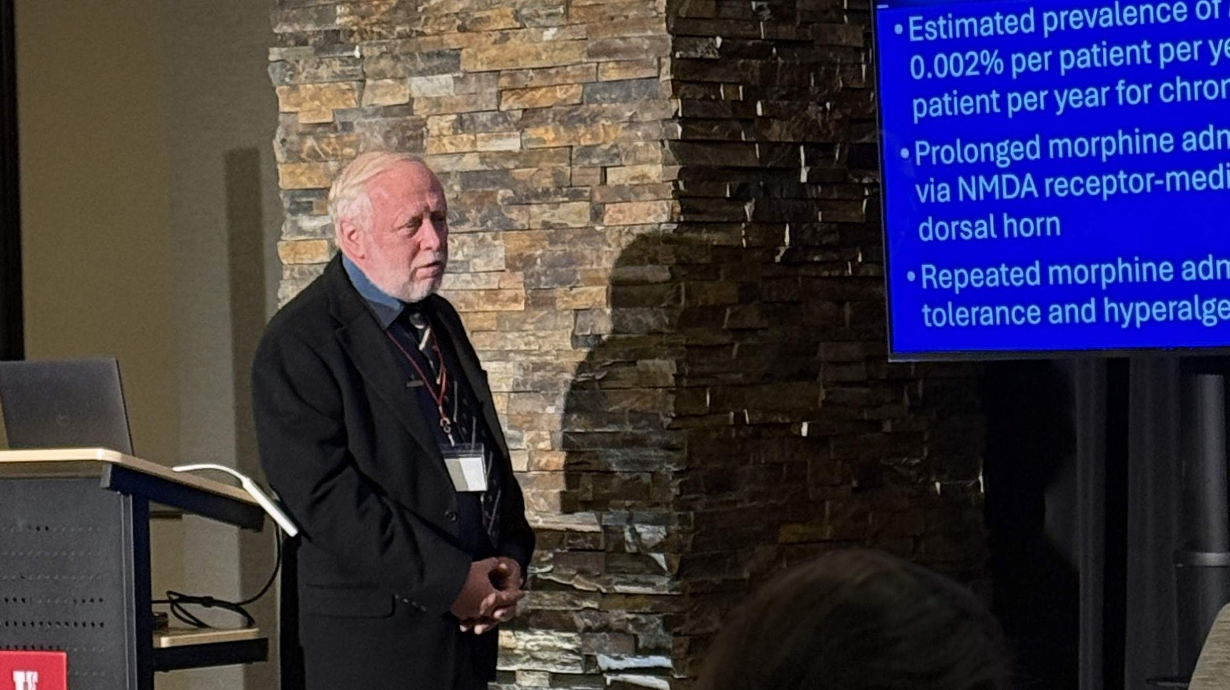 Dr. Alexander A Vitin presents at the postgrad conference in a dark blue suit, standing at a podium and standing in front of a screen displaying slides from his presentation.