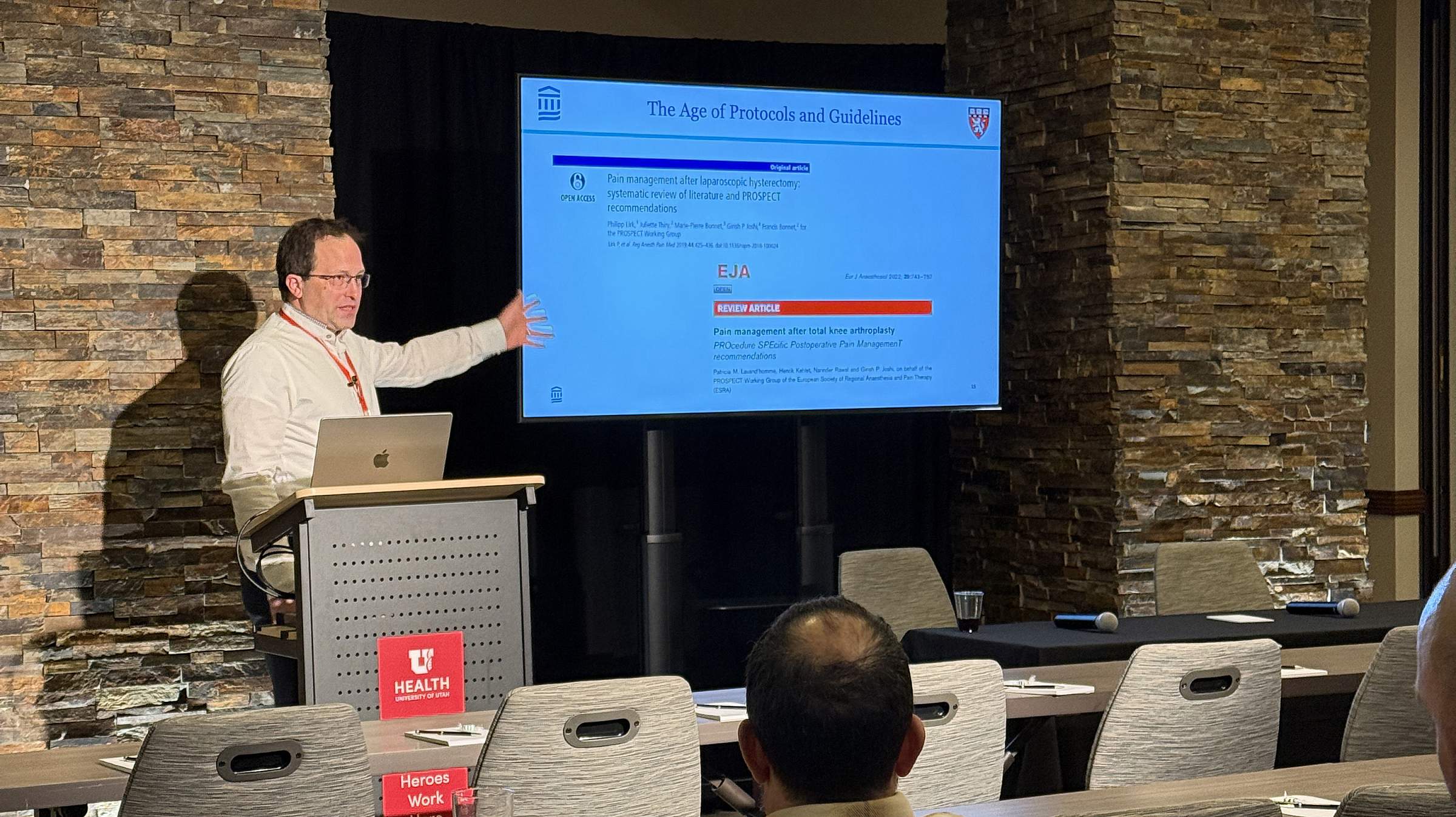 Dr. Philipp Lirk presents at a postgrad conference. He stands at a podium in front of a screen, wearing a white button-down shirt with a red lanyard.