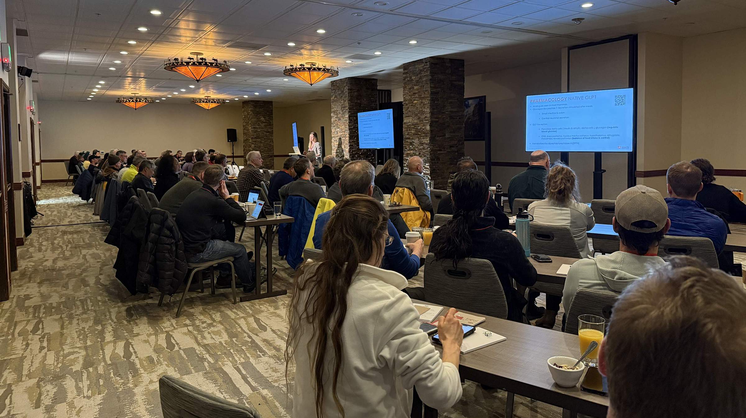 Attendees gather in a ballroom to listen to a speaker. Three screens at the front of the room display the speaker's presentation.