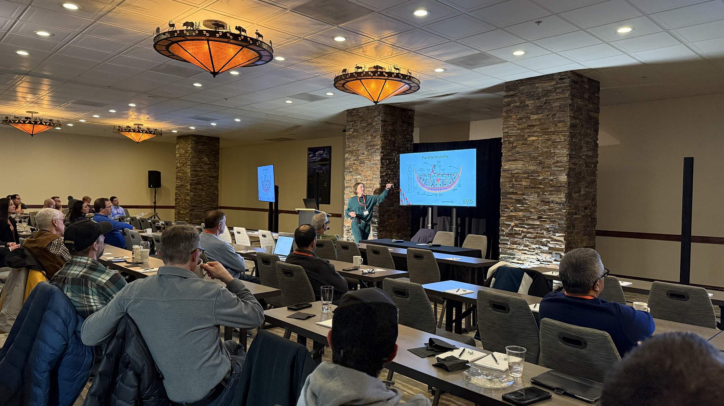 Attendees gather in a ballroom to listen to a speaker. Two screens at the front of the room display the speaker's presentation.