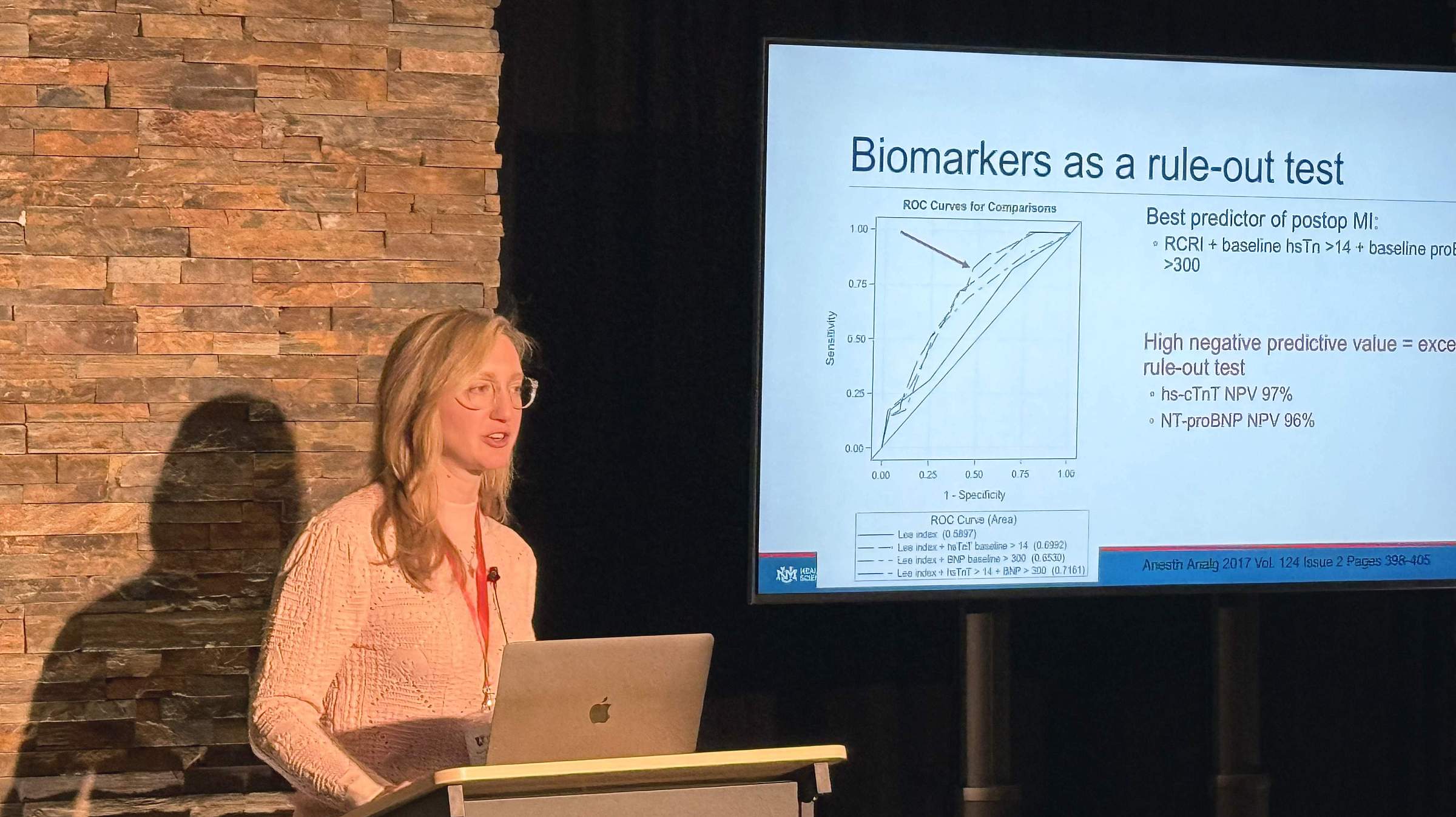 Dr. Emilee Borgmeier presents at the postgrad conference. She stands at a podium in front of a screen. She wears a pink button-down shirt and glasses.