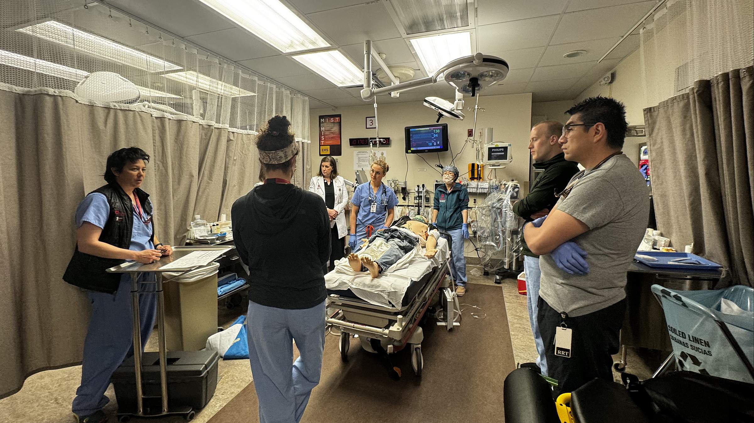 Participants in an in-situ simulation at the Center for Patient Simulation. There are seven people gathered around a simulated patient on a gurney. The room they are standing in is divided by a grey privacy screen.